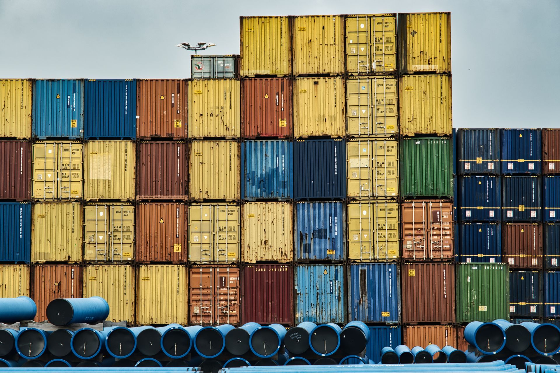 Stacked colorful shipping containers in a cargo yard with blue barrels in front