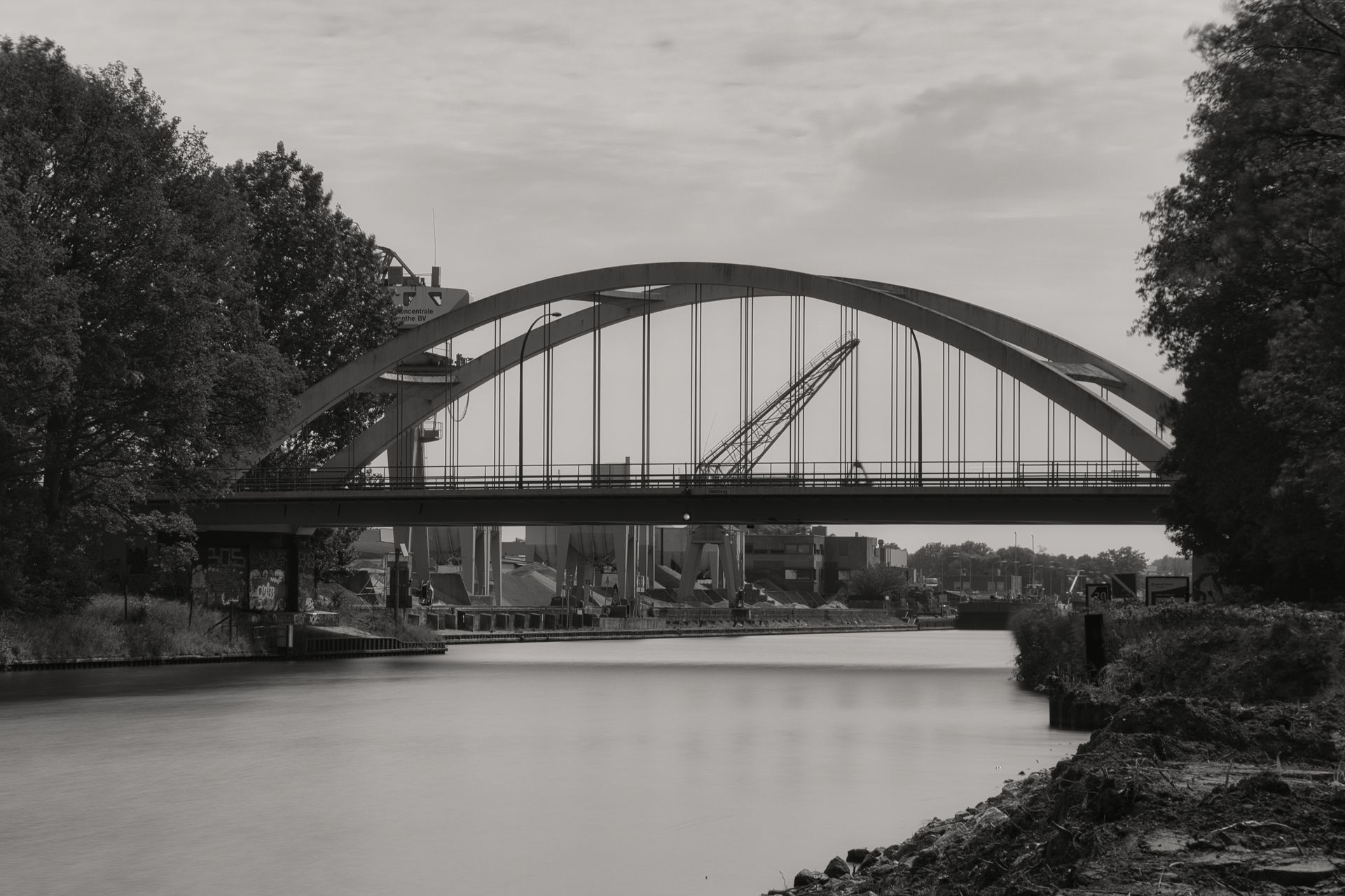 Black-and-white view of an arched bridge spanning a calm river, framed by trees and a cloudy sky