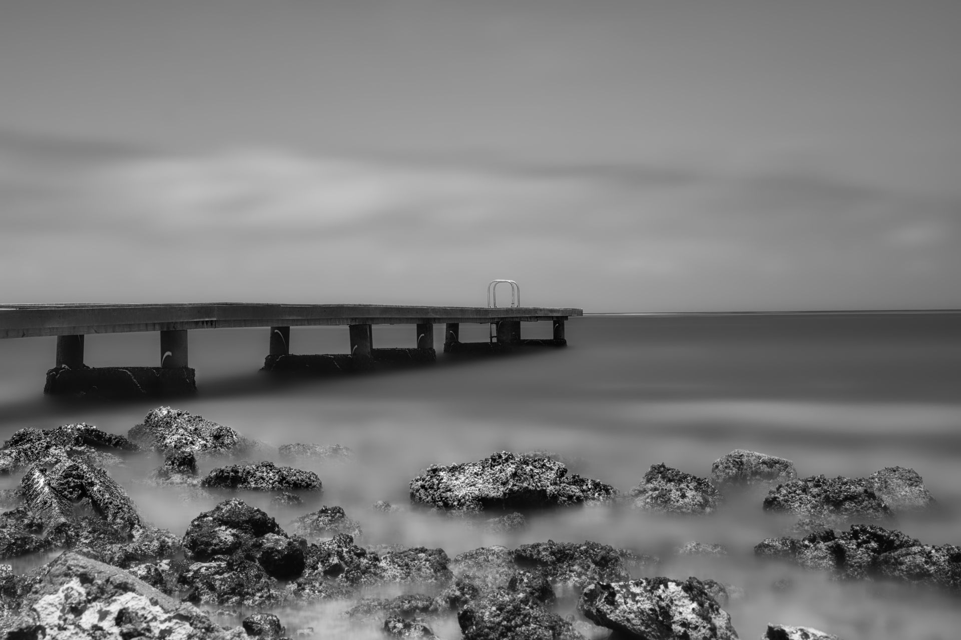 Black-and-white seascape with a long pier over calm water and rocky shore in the foreground