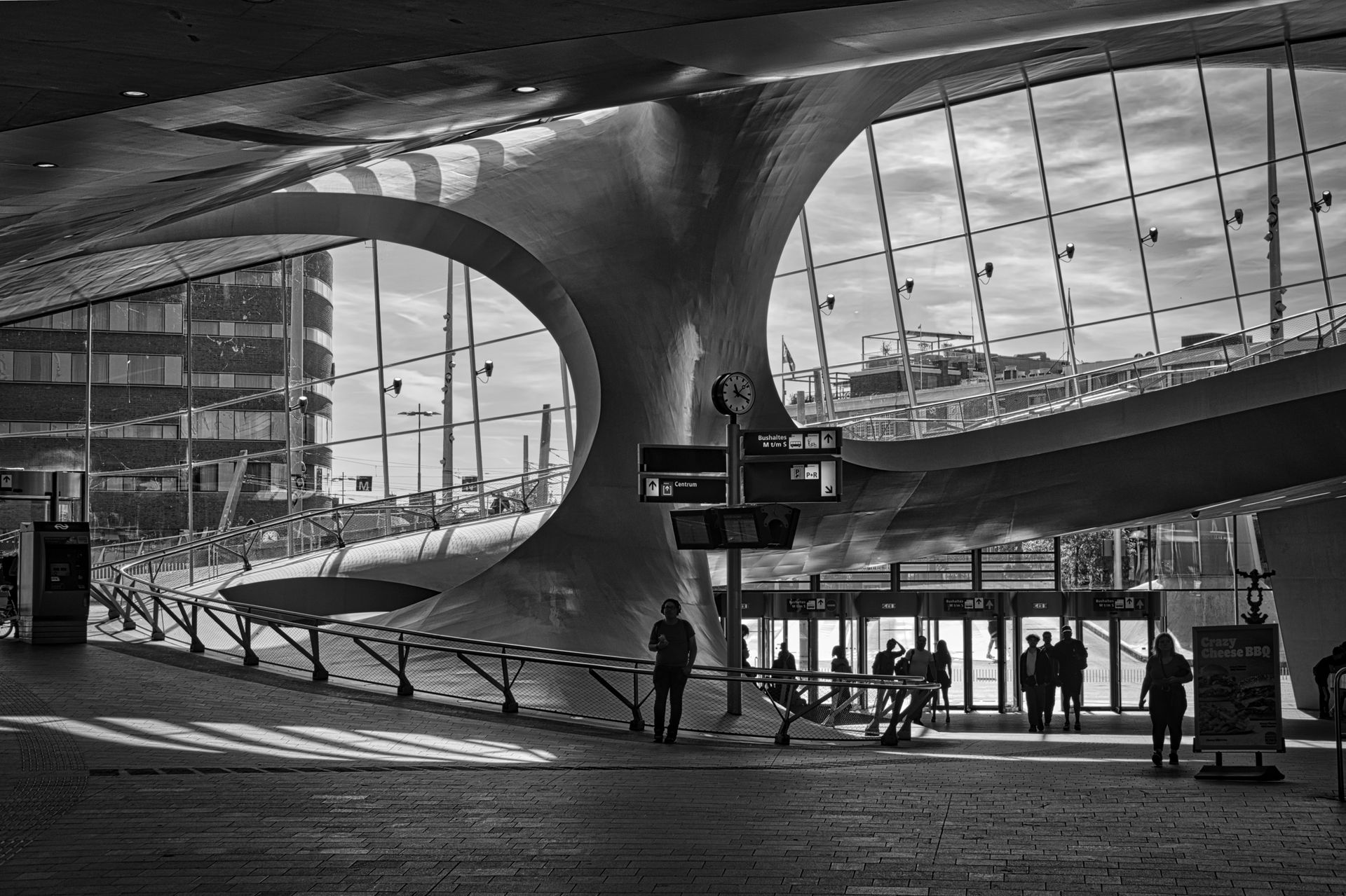 Black-and-white railway station terminal with curved glass roof, waiting travelers, and departure sign at dusk