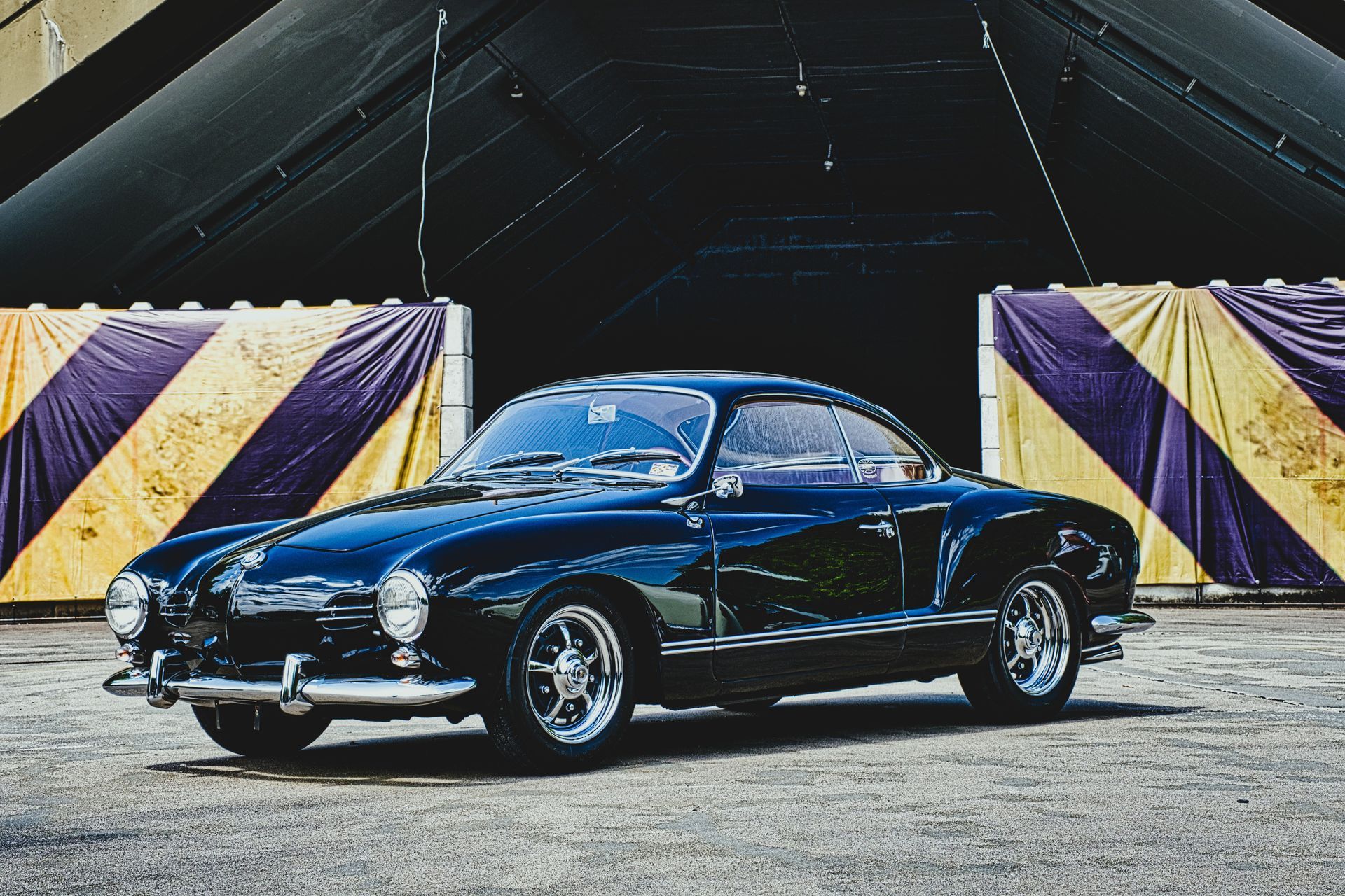 Black Karmann Ghia parked before purple and yellow draped tents on a gravel lot