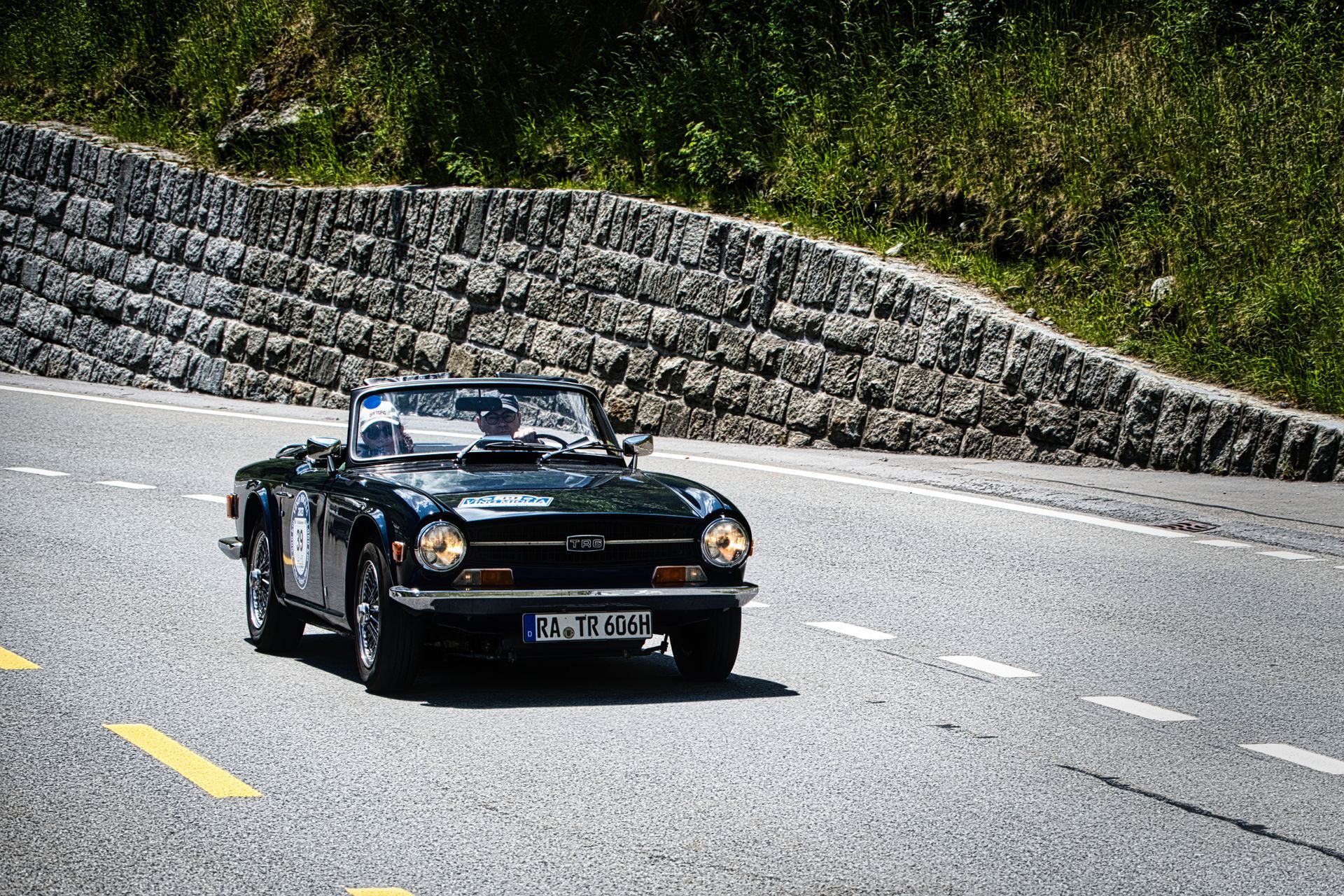 Triumph TR6 driving on a curving road beside a stone wall and greenery