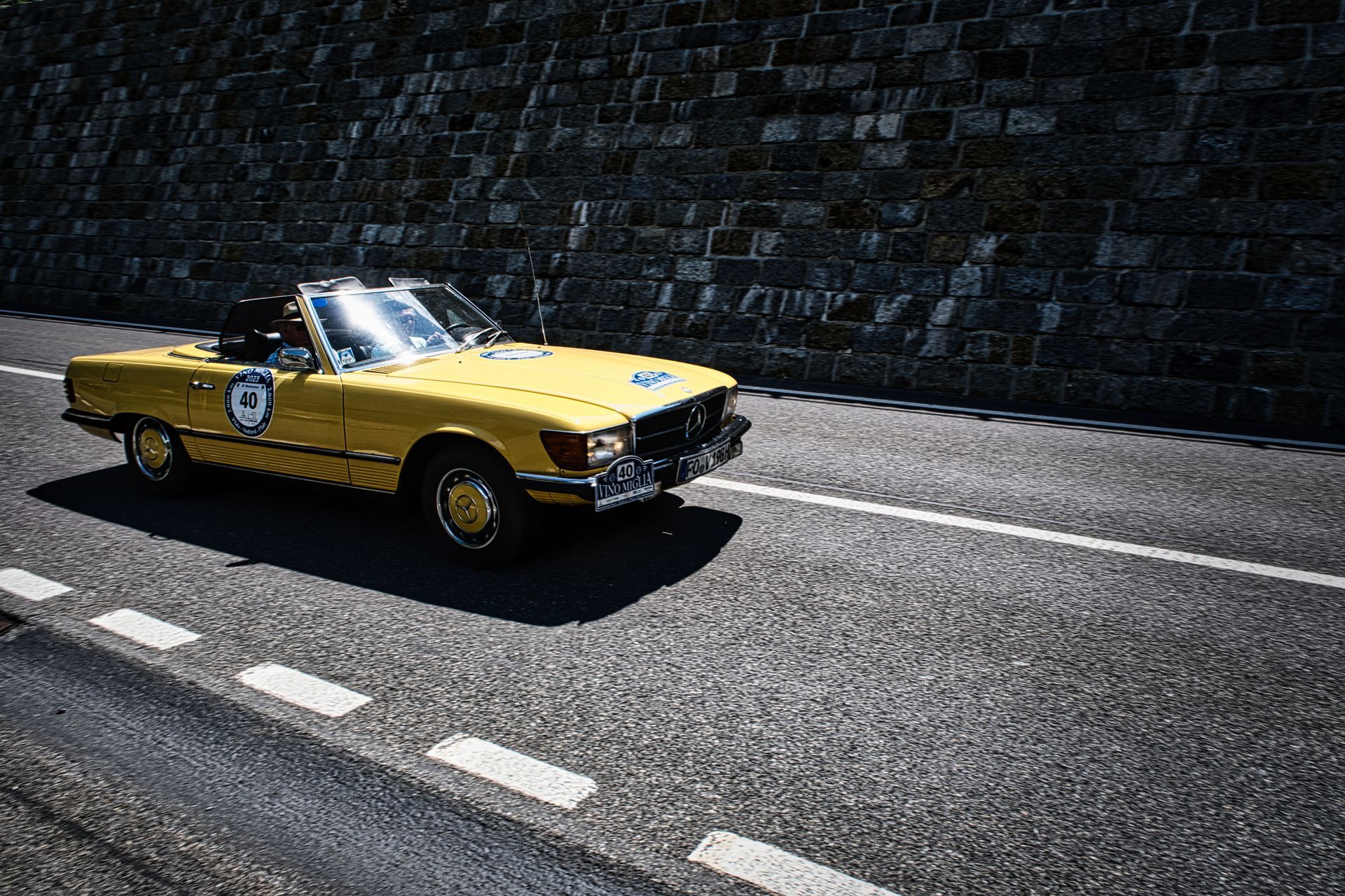 Mercedes 350 SL vintage convertible driving on a road beside a dark stone wall.