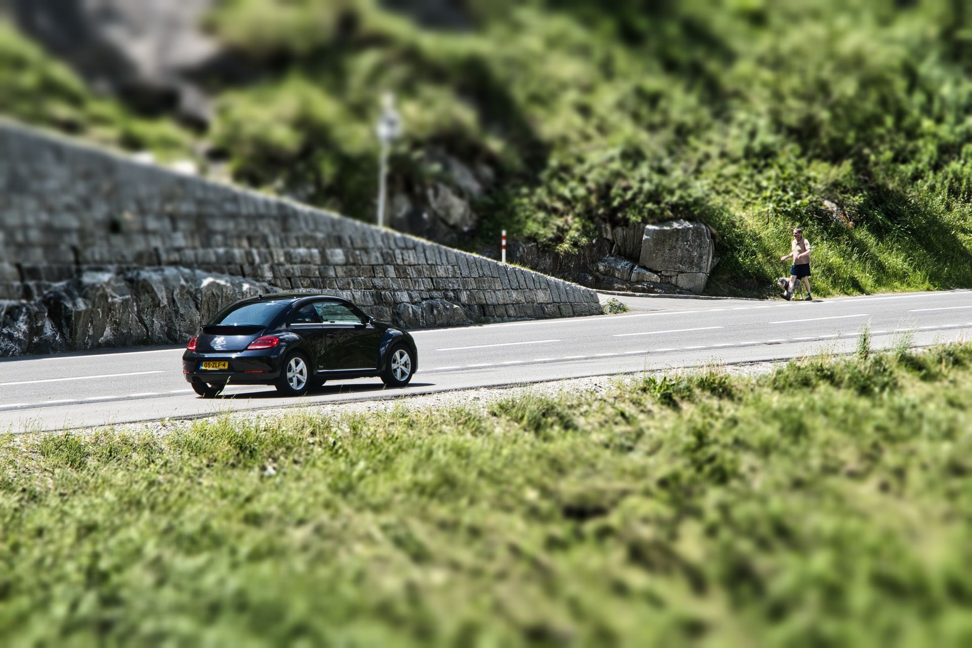 Volkswage Beetle driving on a winding mountain road beside a stone wall and green hills.