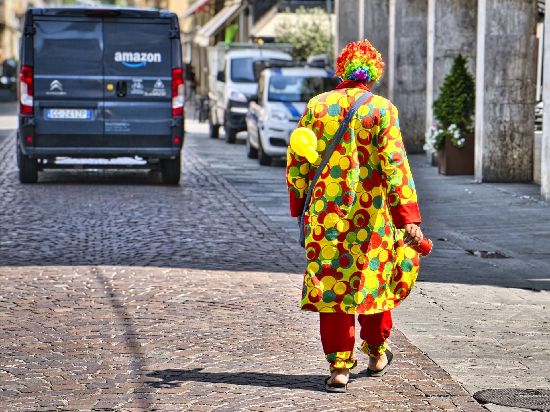 Person in a bright floral outfit walking down a cobblestone street behind an Amazon van