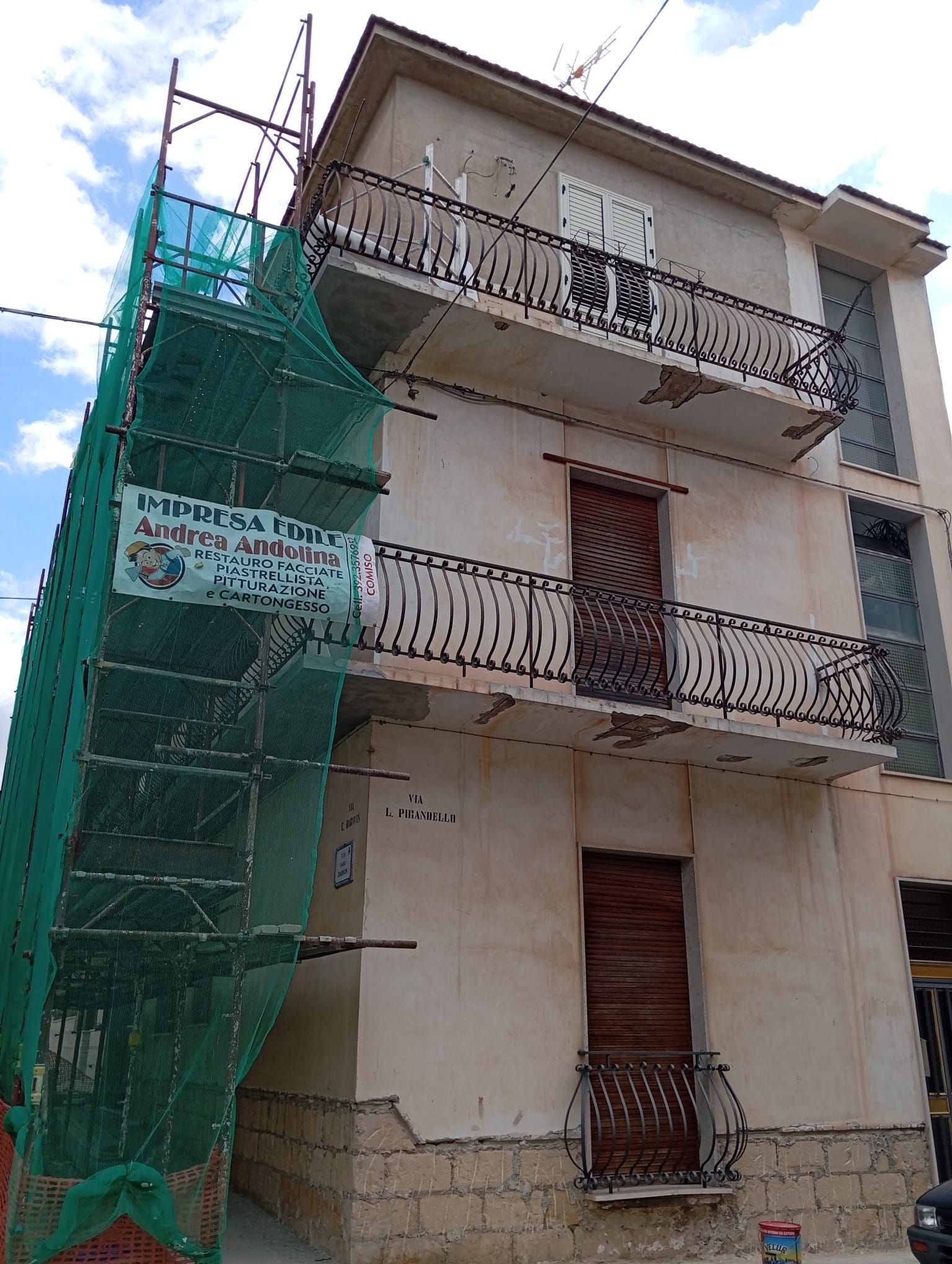 Edificio residenziale di tre piani con balconi e rete di ponteggio verde sul lato sinistro.
