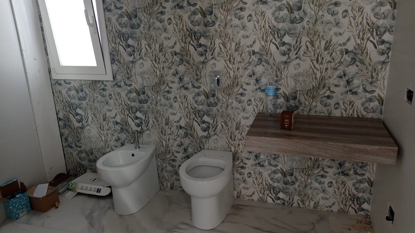 Piccolo bagno con carta da parati a motivi floreali, WC, lavandino, finestra e piano di lavoro in legno con oggetti.