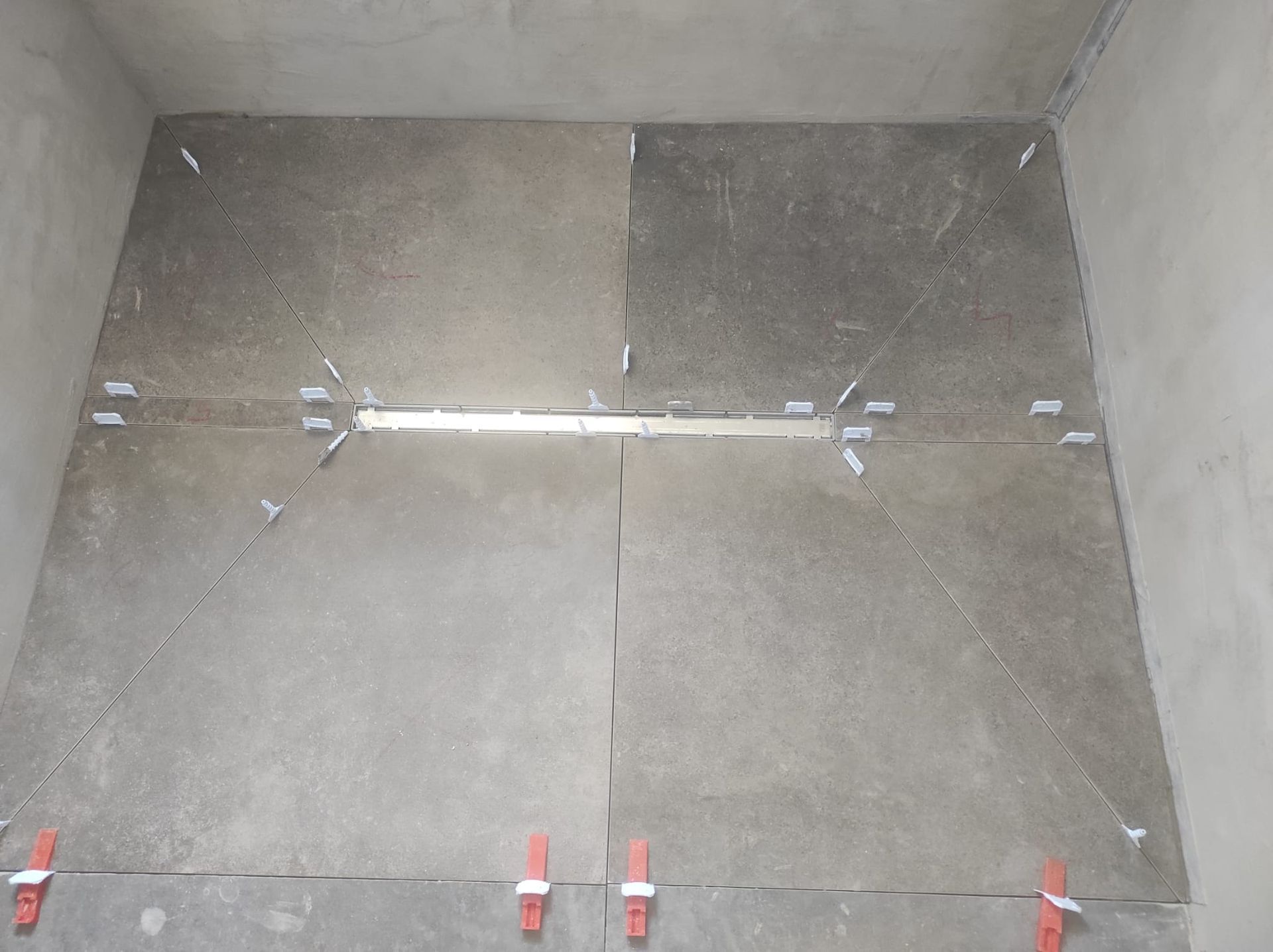 Angolo vuoto di una stanza in cemento con pareti e pavimento grigi, che mostra una giunzione centrale e punti di ancoraggio metallici.
