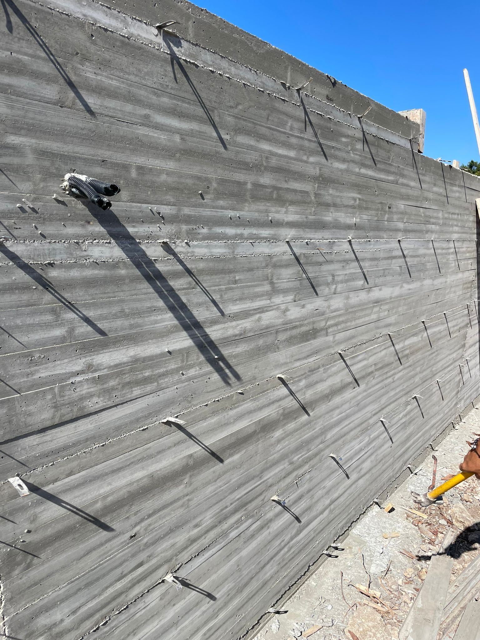 Muro di contenimento in cemento armato in costruzione con ancoraggi in tondino e lunghe ombre sotto la luce intensa del sole.
