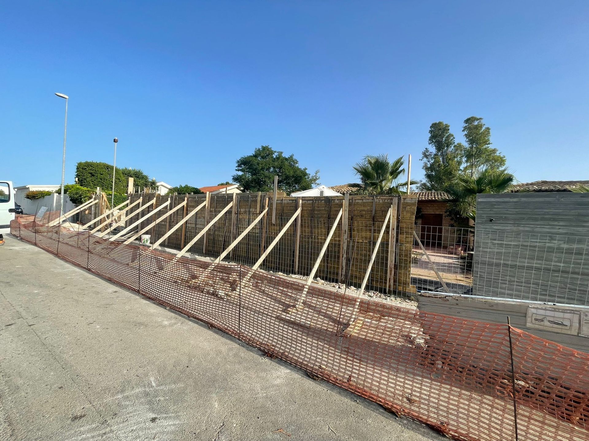 Muro di contenimento in costruzione lungo una strada, con puntelli in legno e recinzione di sicurezza arancione.