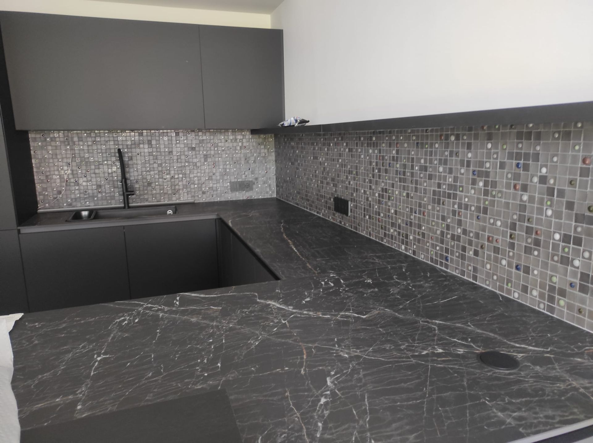 Cucina moderna con mobili scuri, ripiani neri e paraschizzi in piastrelle a mosaico grigie.