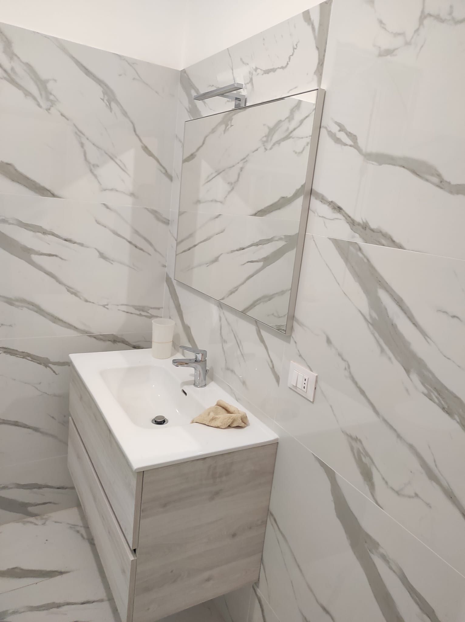 Piccolo bagno con pareti rivestite con piastrelle effetto marmo, mobiletto in legno, lavabo, specchio e portarotolo di carta igienica.