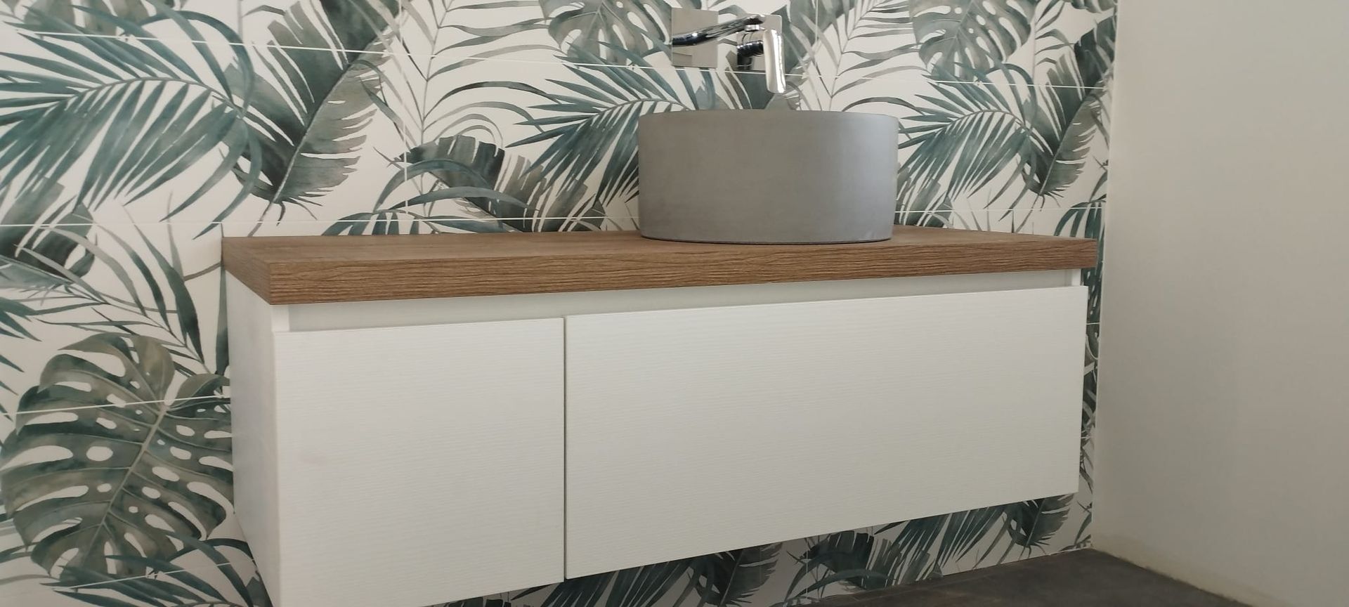 Mobiletto a muro bianco con piano in legno e fioriera grigia su carta da parati a foglie tropicali.