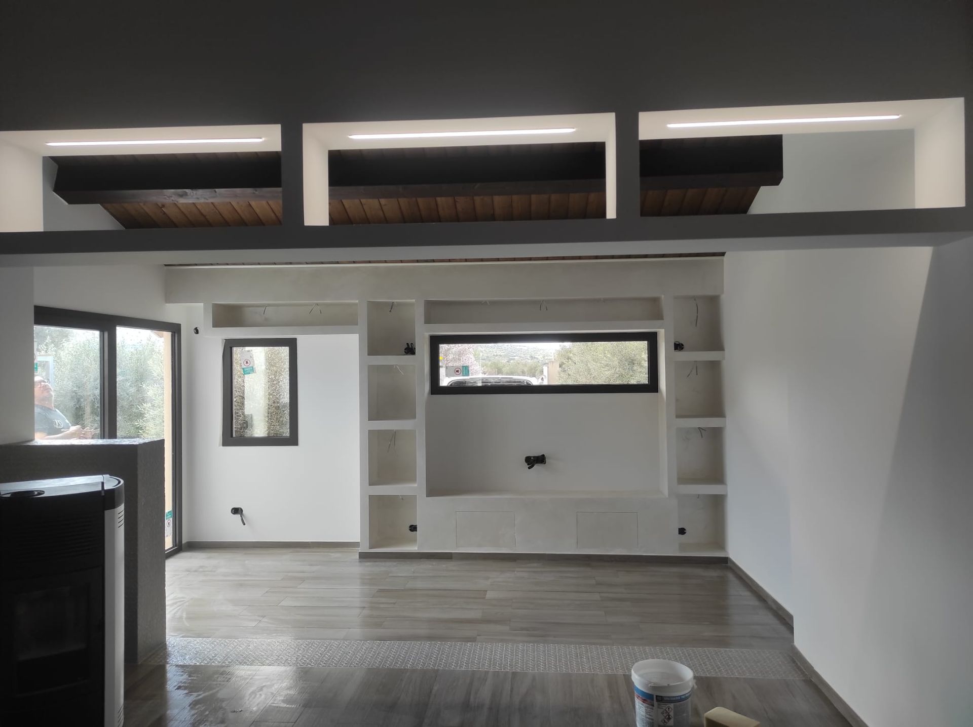 Stanza vuota e non rifinita con scaffali a muro bianchi, finestre alte e travi a vista sul soffitto.