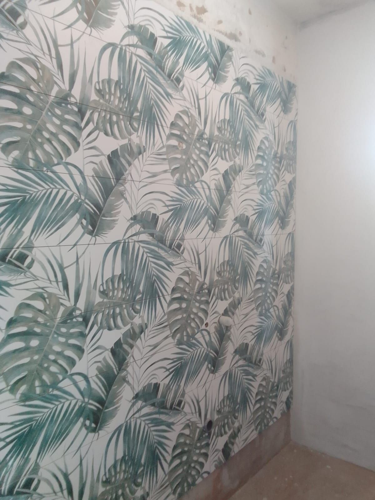 Angolo di una stanza con carta da parati a foglie tropicali verdi e una parete bianca semplice.