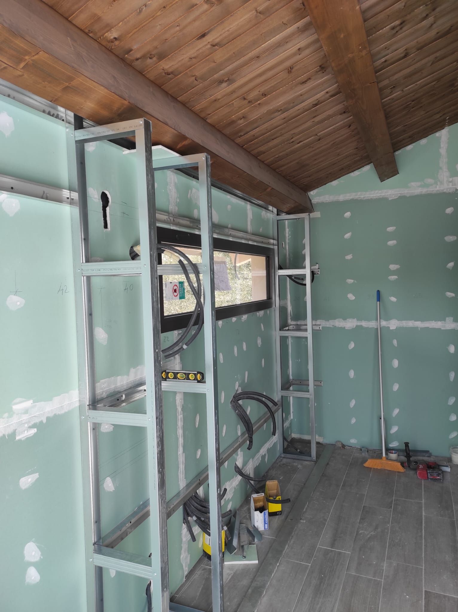 Stanza parzialmente rifinita con pareti in cartongesso verde, soffitto con travi a vista e attrezzi da costruzione sul pavimento.