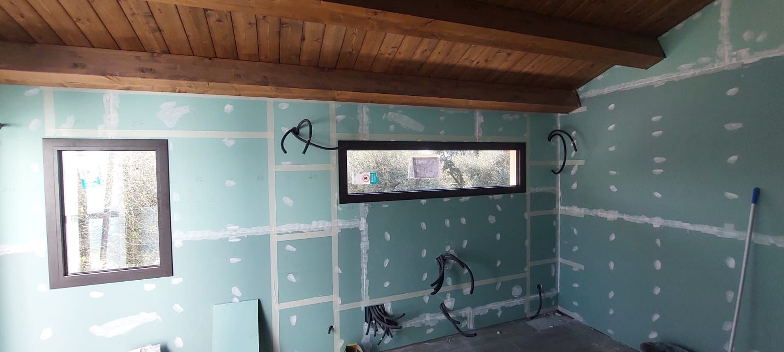 Interno di una stanza color verde acqua non rifinito, con cavi elettrici a vista, soffitto in legno e due finestre strette.
