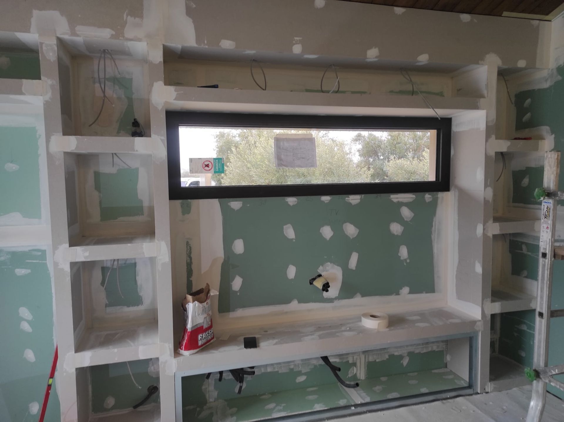 Stanza parzialmente ristrutturata con pareti in cartongesso verdi, stuccature bianche e una lunga finestra sopra una mensola a muro.