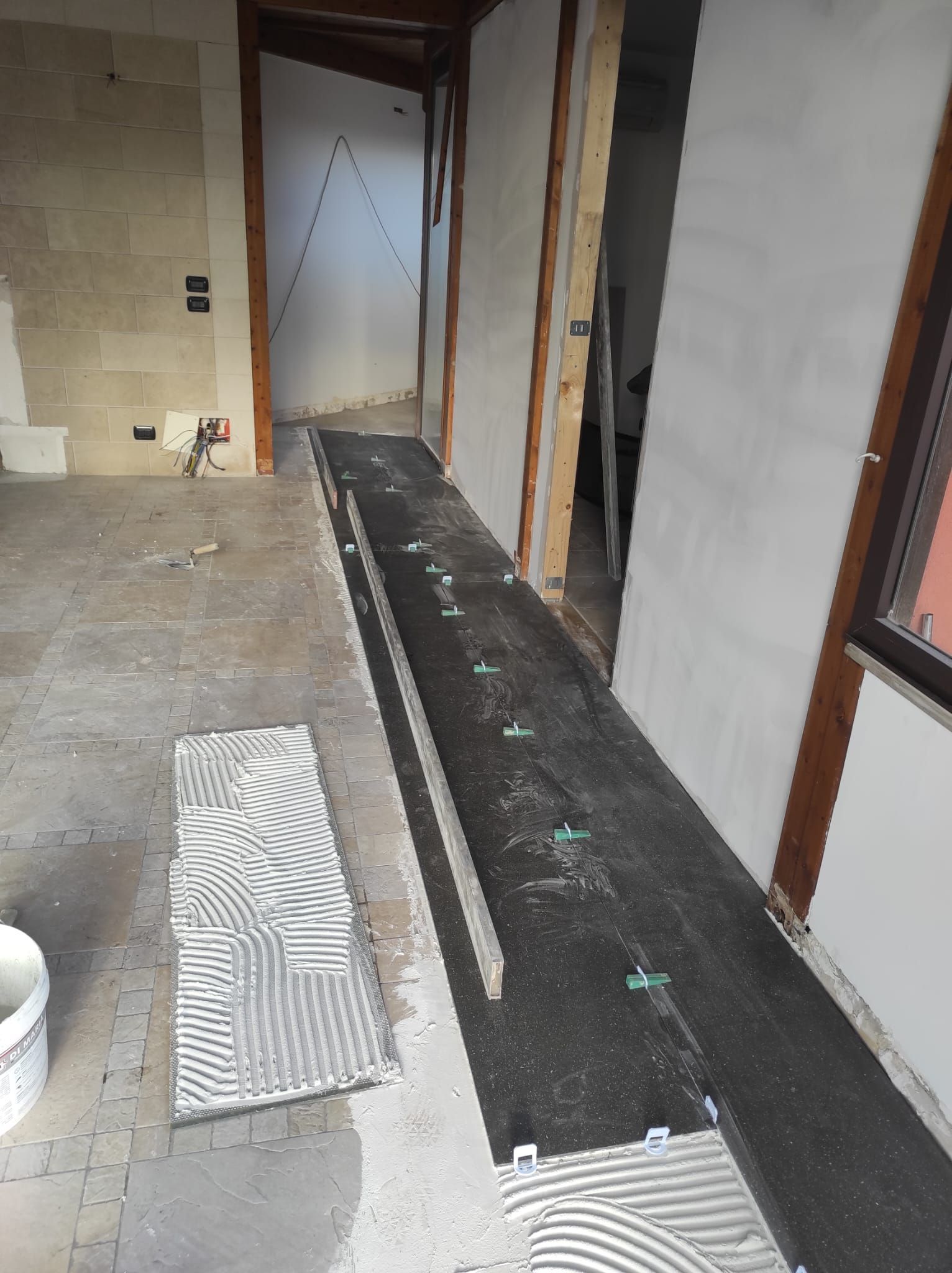 Lungo e stretto corridoio in costruzione con pareti a vista, pavimento in membrana nera e una porta in fondo.