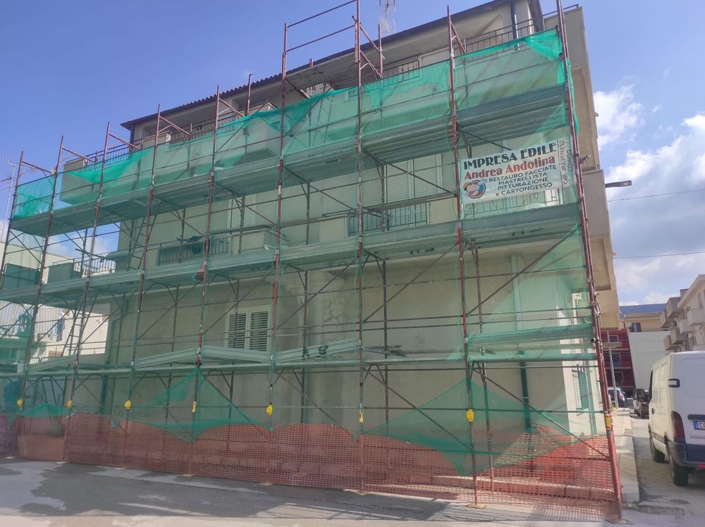 Edificio in ristrutturazione avvolto da una rete di impalcature verdi, accanto a una strada sotto un cielo azzurro.