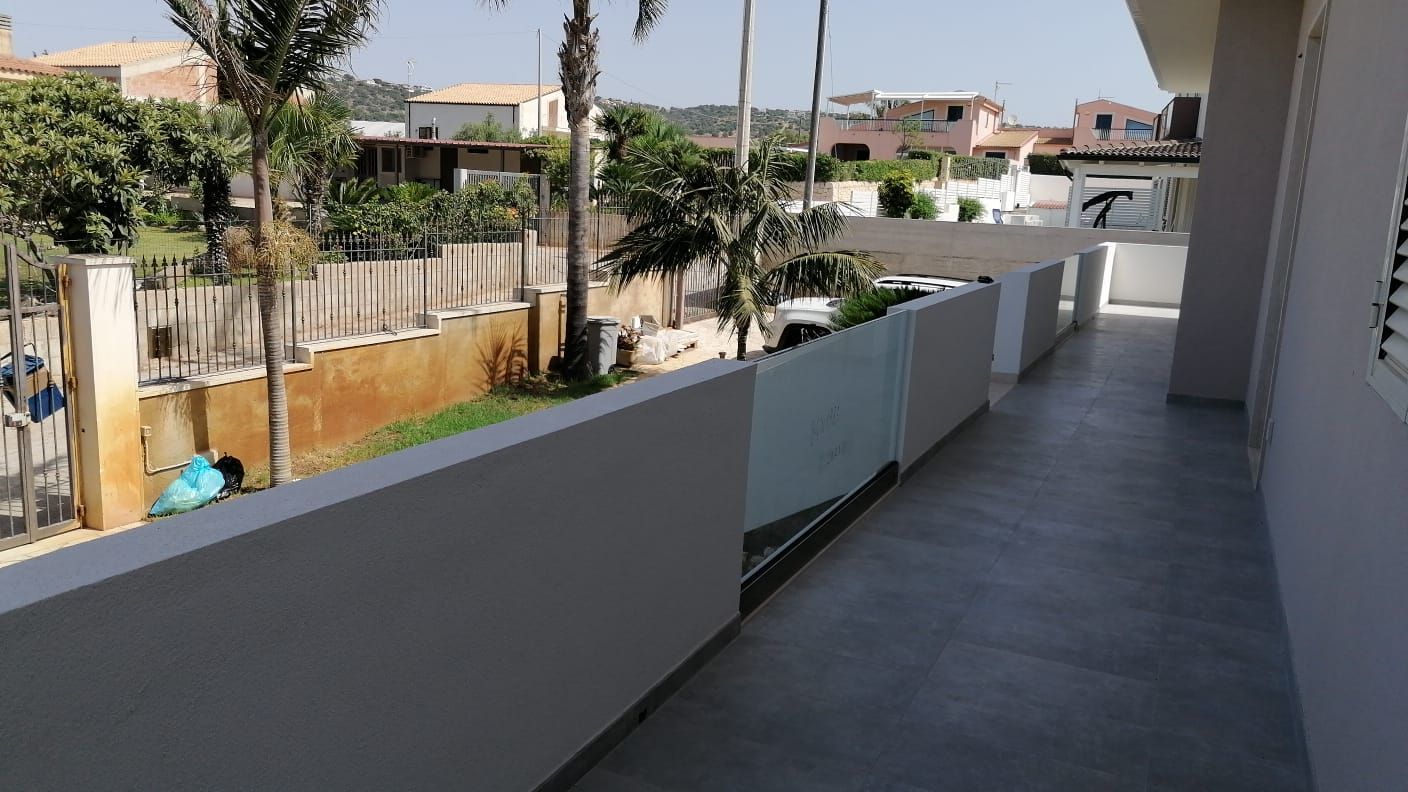 Passerella del balcone con parete grigia, affacciata su una strada del quartiere e palme sotto un sole splendente