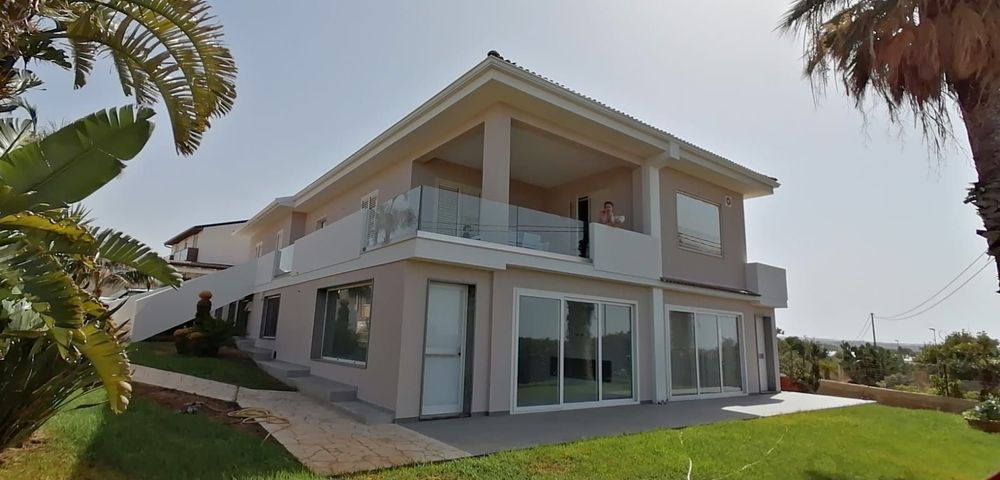 Casa moderna a due piani di colore bianco con ampie finestre, balcone in vetro e prato verde sotto le palme.