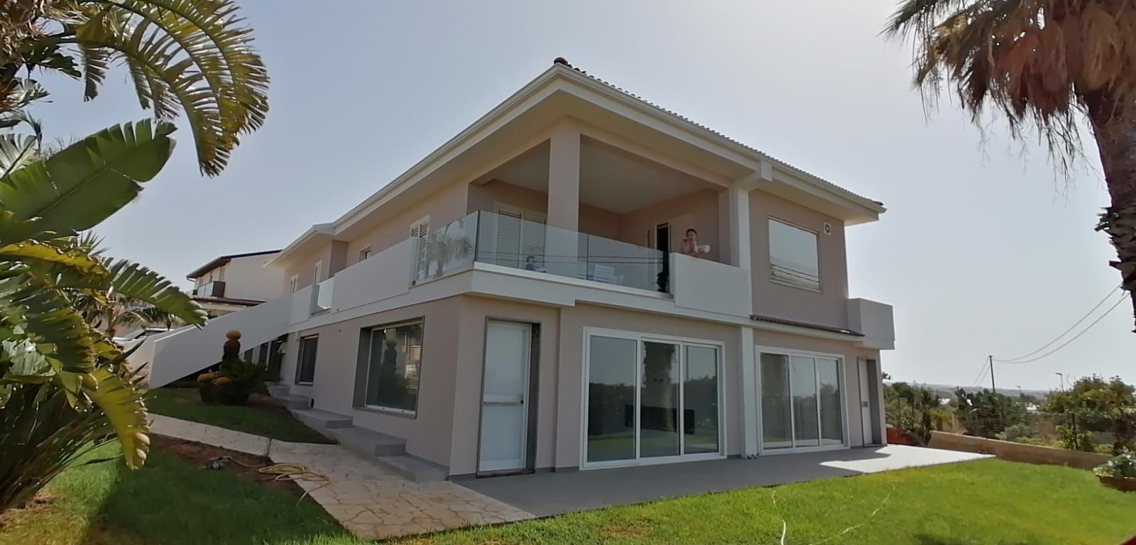 Casa moderna a due piani di colore bianco con balconi in vetro e ampie finestre, immersa in un soleggiato giardino paesaggistico.