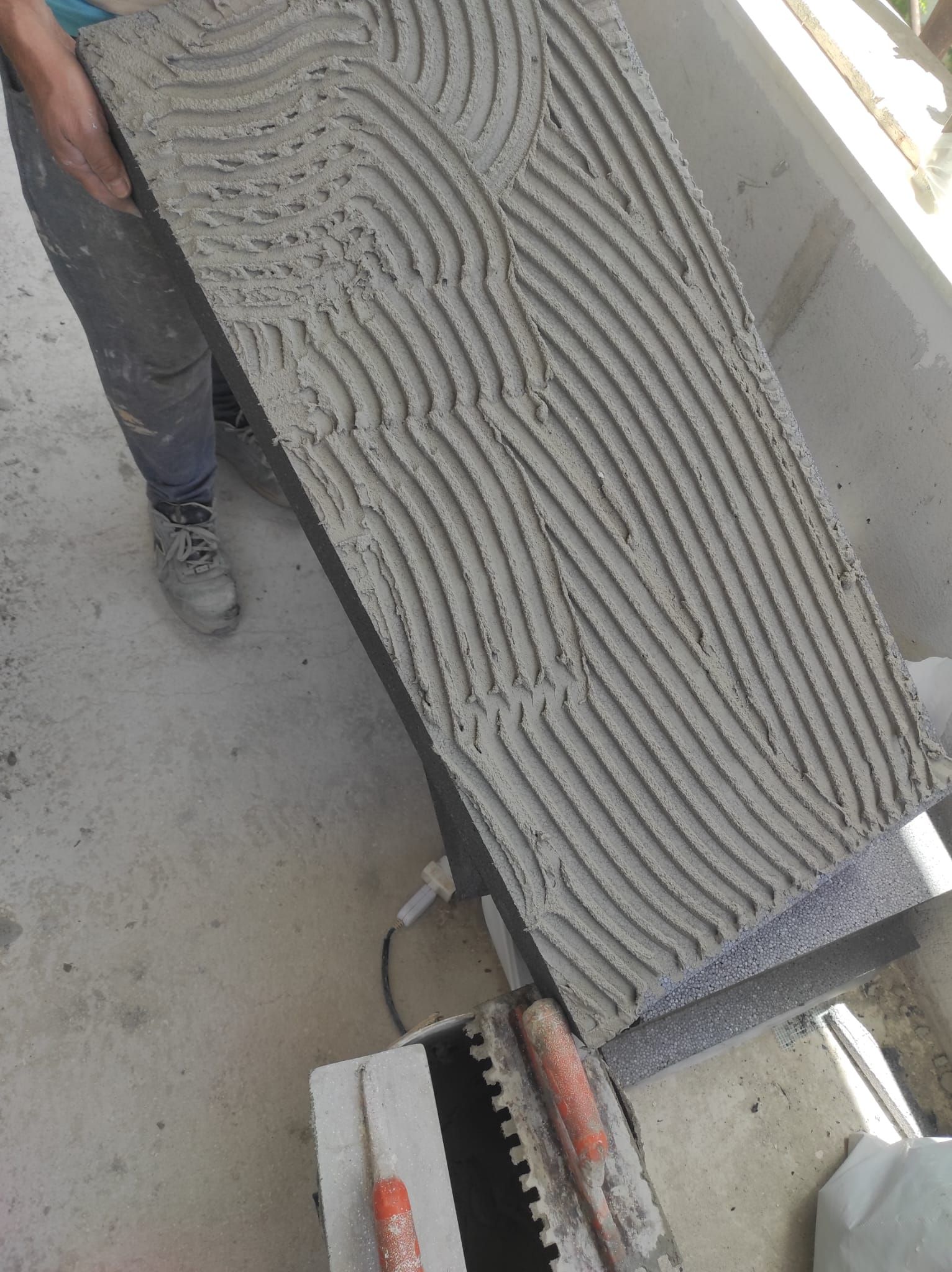 Primo piano di una grande lastra di cemento con superficie ruvida, maneggiata accanto alla gamba di una persona e a un utensile elettrico.