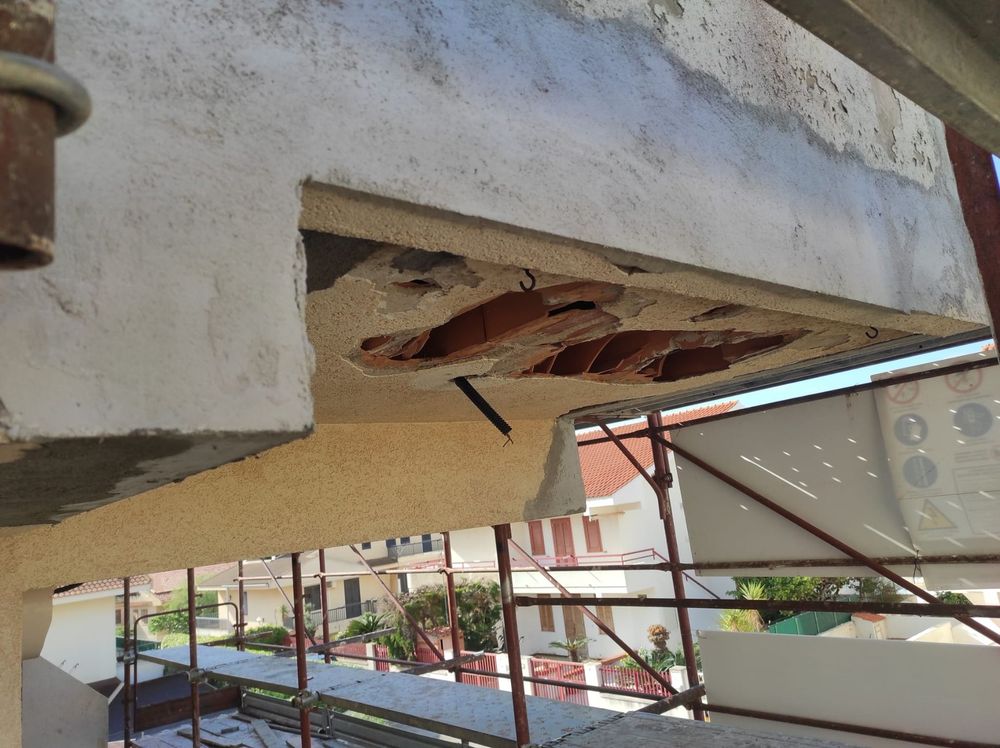 Parte inferiore di un balcone in cemento rotto, con barre d'armatura a vista e intonaco scheggiato sopra una ringhiera.