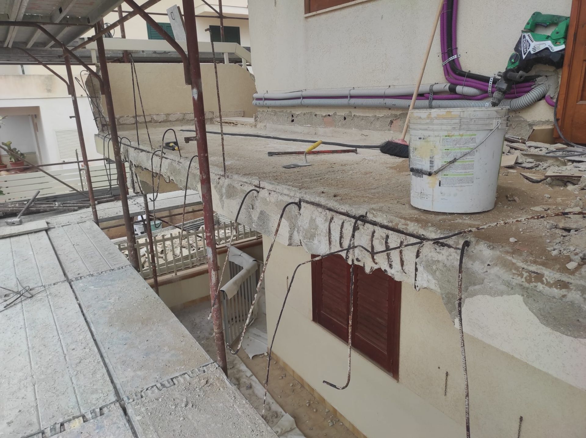 Scena di un cantiere sul tetto con cemento crepato, tubi e un secchio bianco vicino all'apertura di una scala.