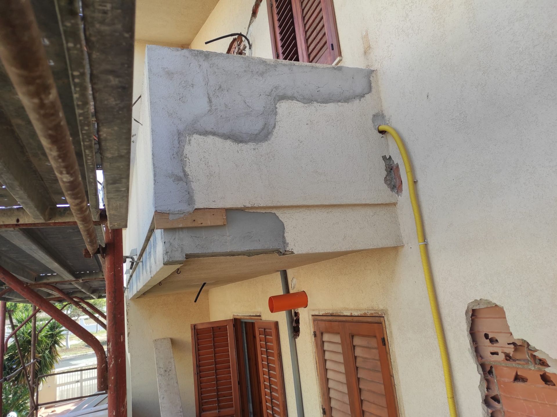 Muro esterno danneggiato con intonaco scrostato sopra le finestre e un tubo giallo sulla facciata di un edificio.