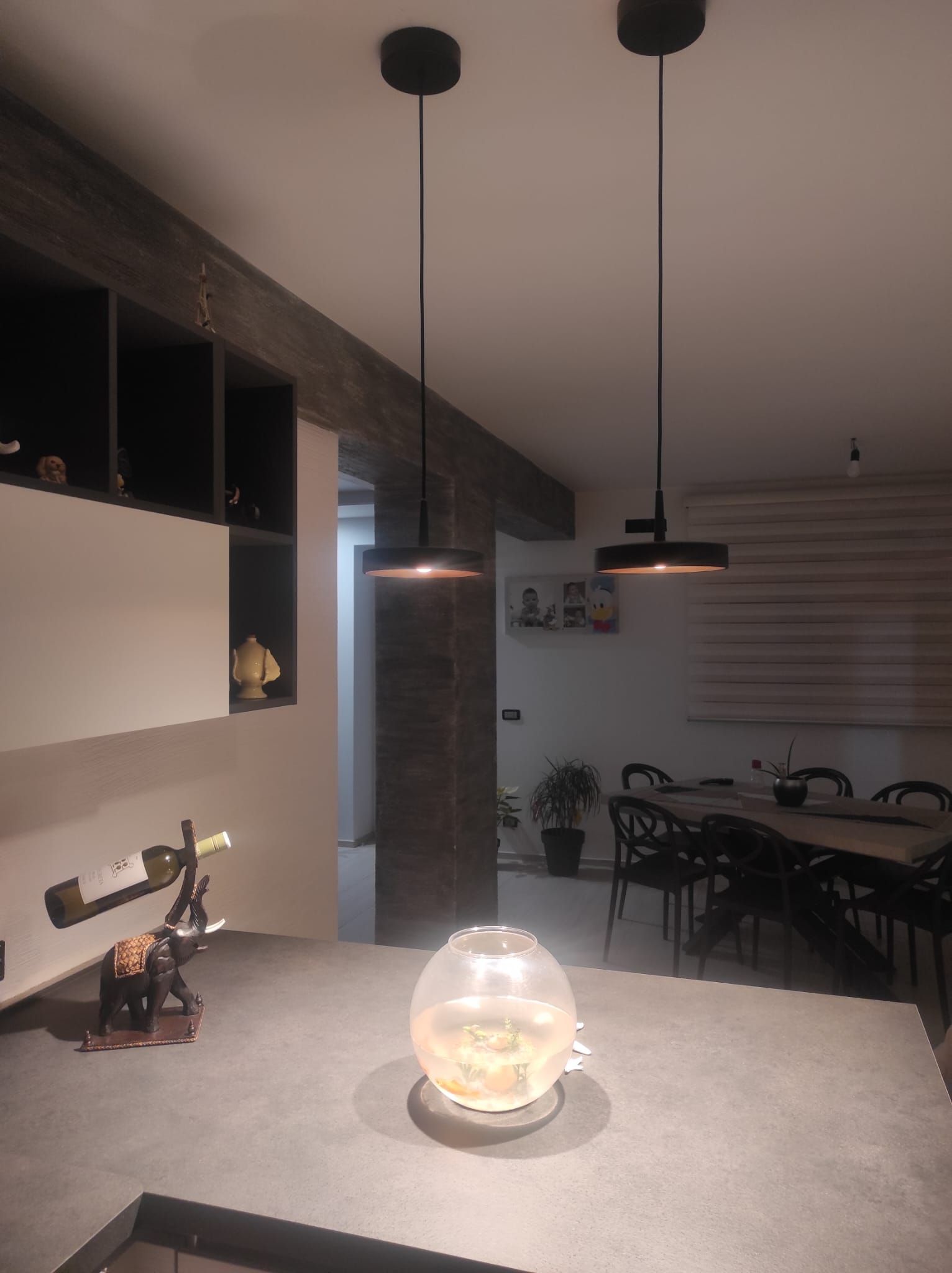 Sala da pranzo moderna con un lungo tavolo, lampade a sospensione e un centrotavola con ciotola di vetro su un bancone.