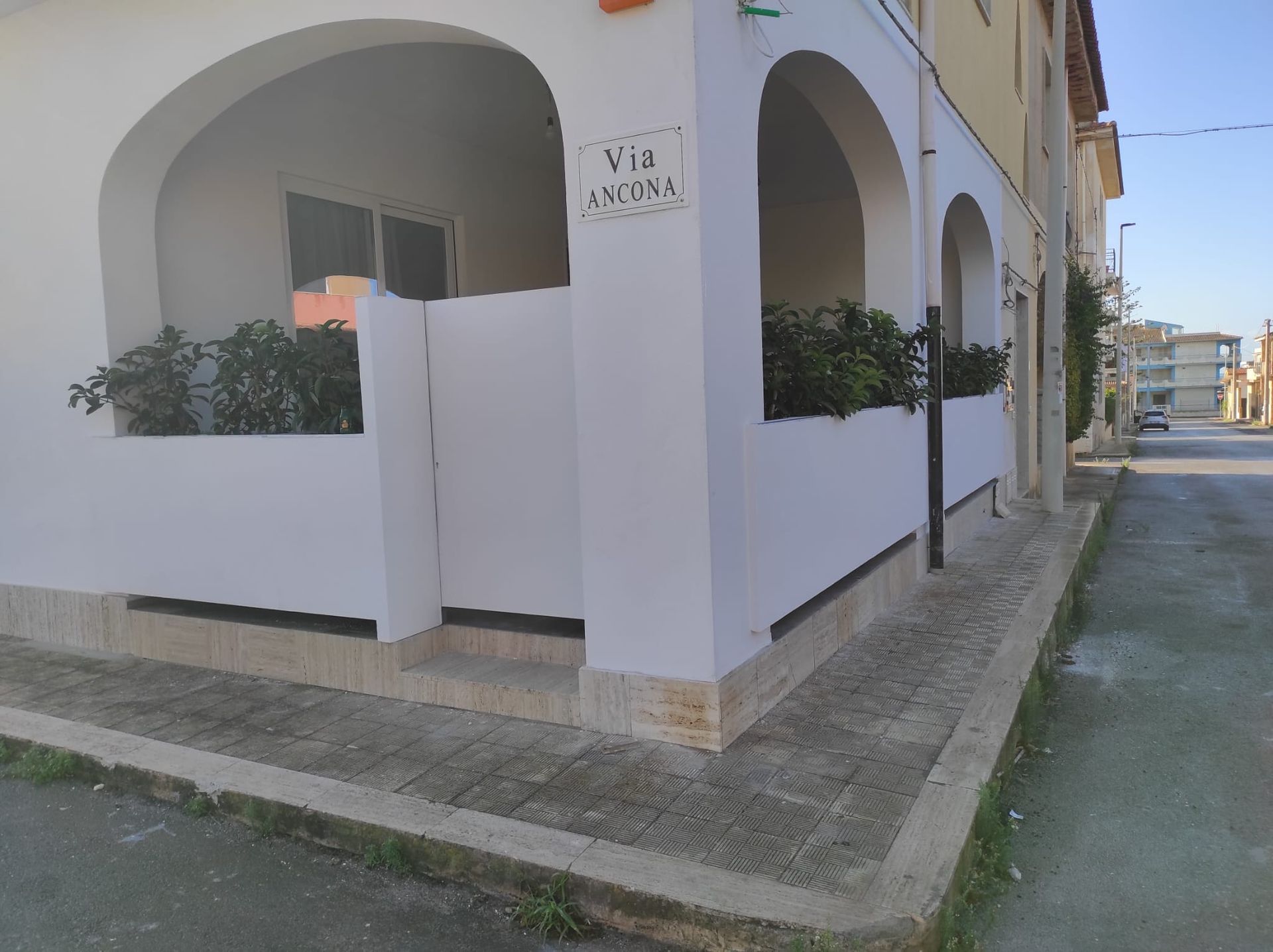 Edificio angolare bianco con portici ad arco e muretto basso, adiacente a una strada tranquilla e a un marciapiede.