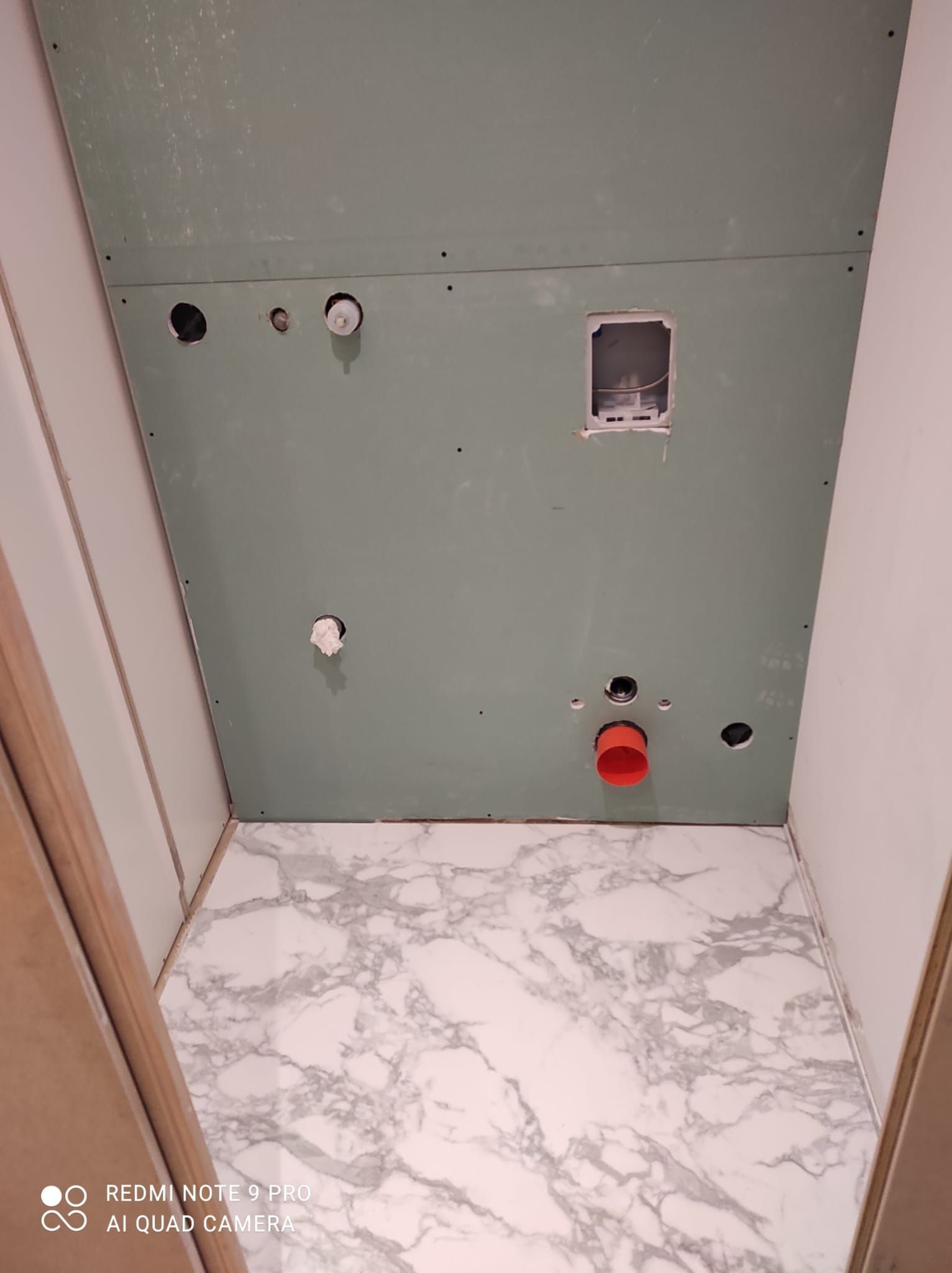 Pannello a parete verde con diversi fori, una presa elettrica rossa e un pavimento con motivo effetto marmo.