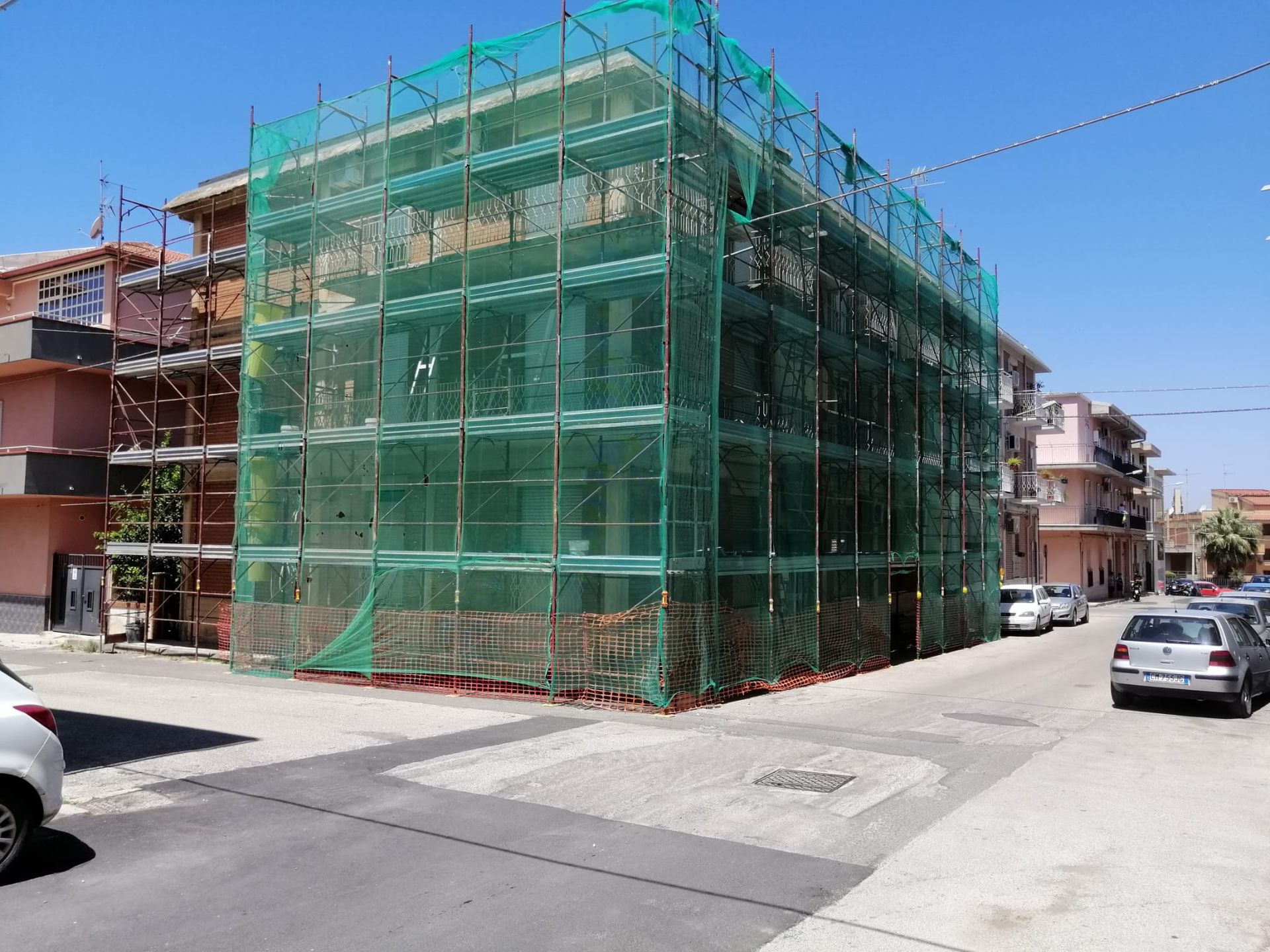 Ponteggi e strutture avvolte in reti verdi in un angolo di strada soleggiato con auto parcheggiate.