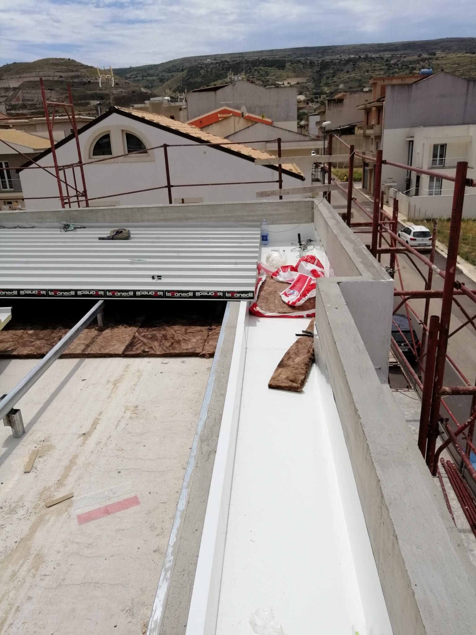Scena di un cantiere su un tetto con pannelli bianchi, travi a vista e una borsa rossa e bianca vicino a muri non finiti.