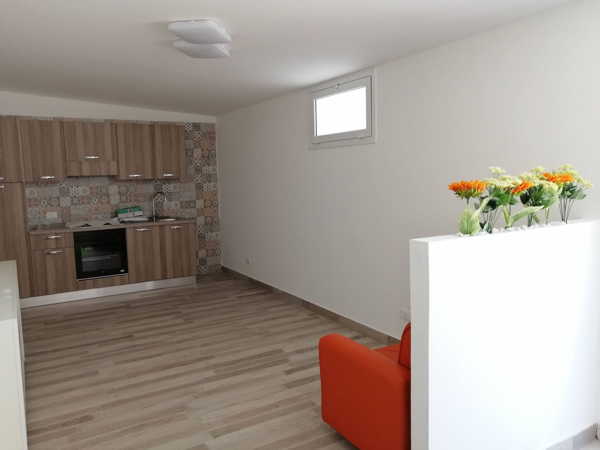 Interno di un appartamento moderno con cucina, divano arancione, pavimento in legno e fiori su un muretto.