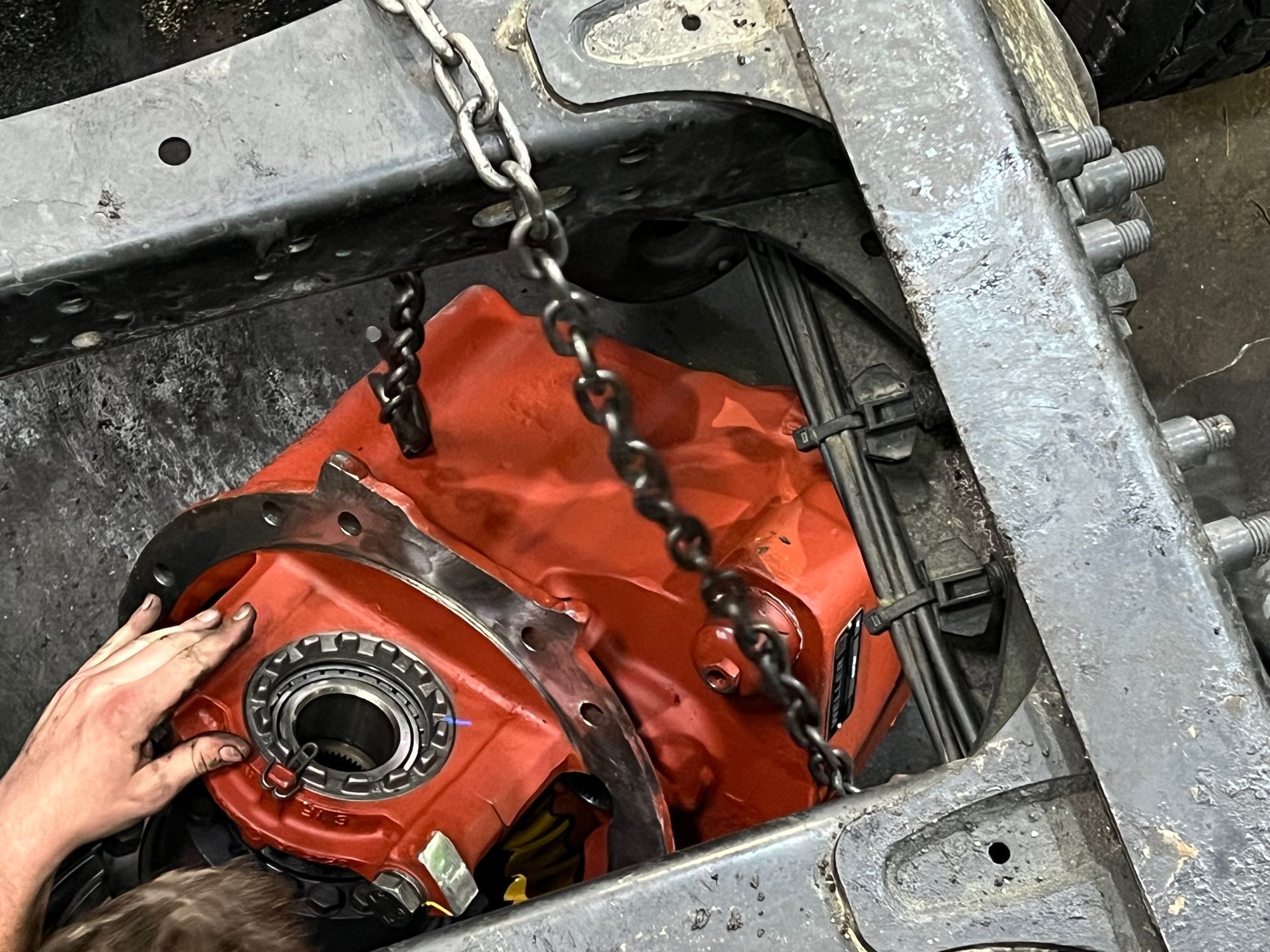 Red gearbox lifted by chains inside a truck frame. A person's hand is on the gearbox.