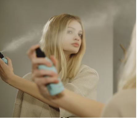 Woman spraying Aveda Heat Relief heat protector into her hair while looking in the mirror