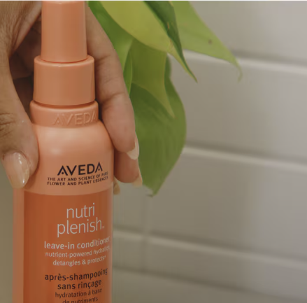Close-up of Aveda Nutriplenish Leave-In Conditioner held in front of green leaves
