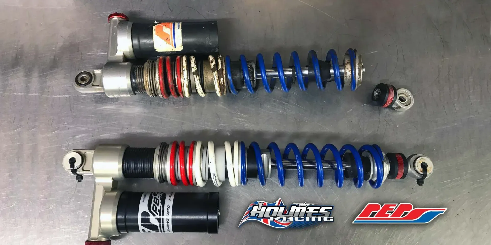 Two high-performance off-road vehicle shocks, one damaged and one intact, displaying red, white, and blue spring coils.