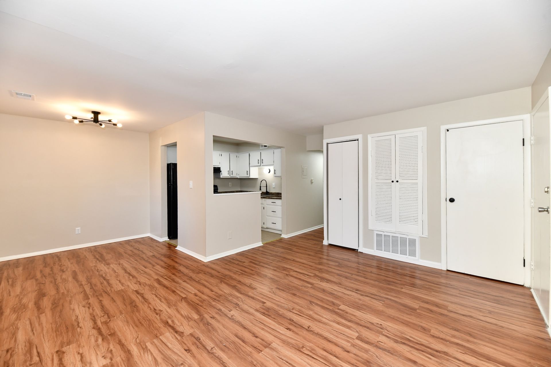 Empty apartment living space with wood flooring, neutral walls, and open kitchen.