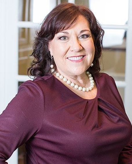 A woman wearing a purple shirt and a pearl necklace is smiling for the camera.