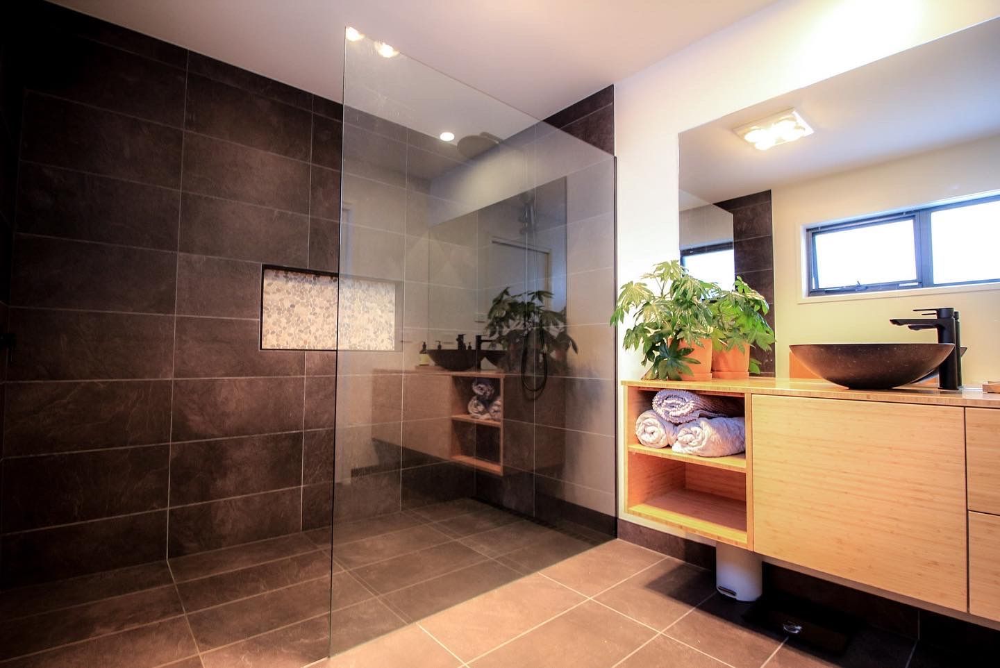 Modern bathroom with a glass-enclosed shower, dark tile walls, and a light wood vanity topped with a green potted plant.