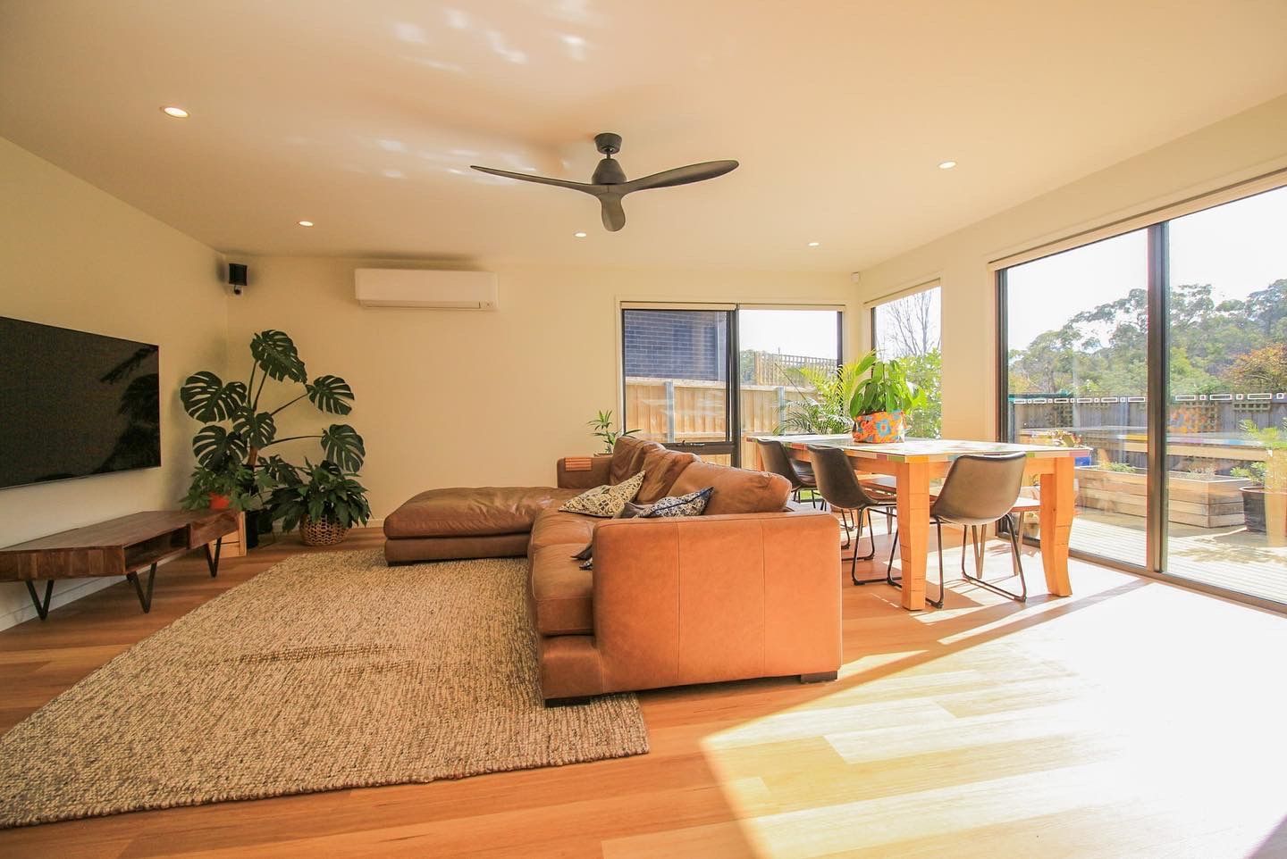 A sunlit living room featuring a tan sectional sofa on a textured rug, a wooden dining set, and a large plant by a TV.