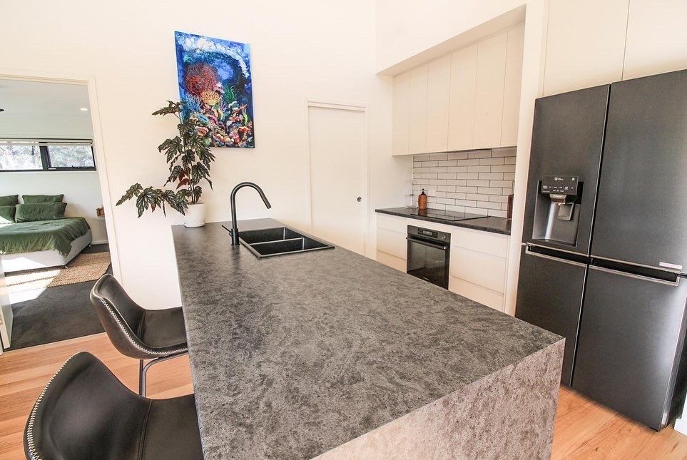 A modern kitchen with a dark grey stone island, black bar stools, stainless appliances, and a black refrigerator.