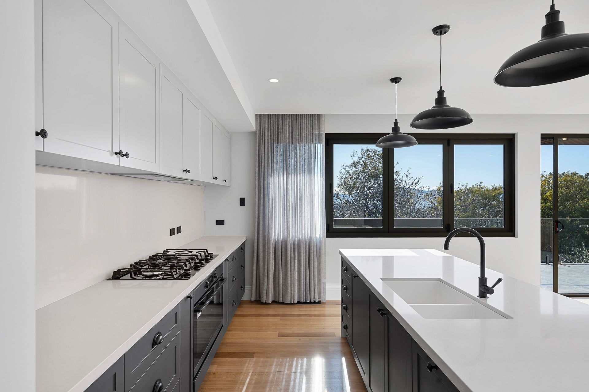 A modern kitchen with dark cabinetry, white countertops, a gas stove, central island, and large windows with grey curtains.