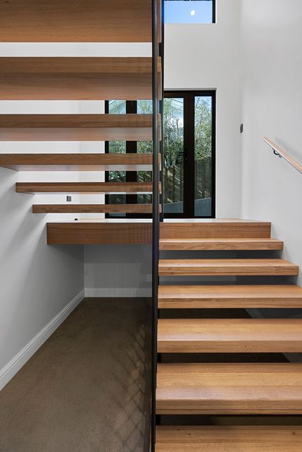 A modern staircase with light wood floating treads and a dark metal railing in a bright white hallway with a window.