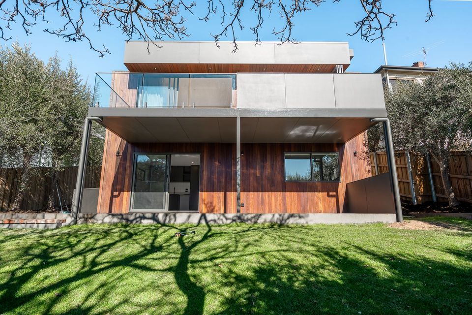 Modern two-story house with wood-paneled walls, a balcony, and sliding glass doors, seen from a green, sunny lawn.