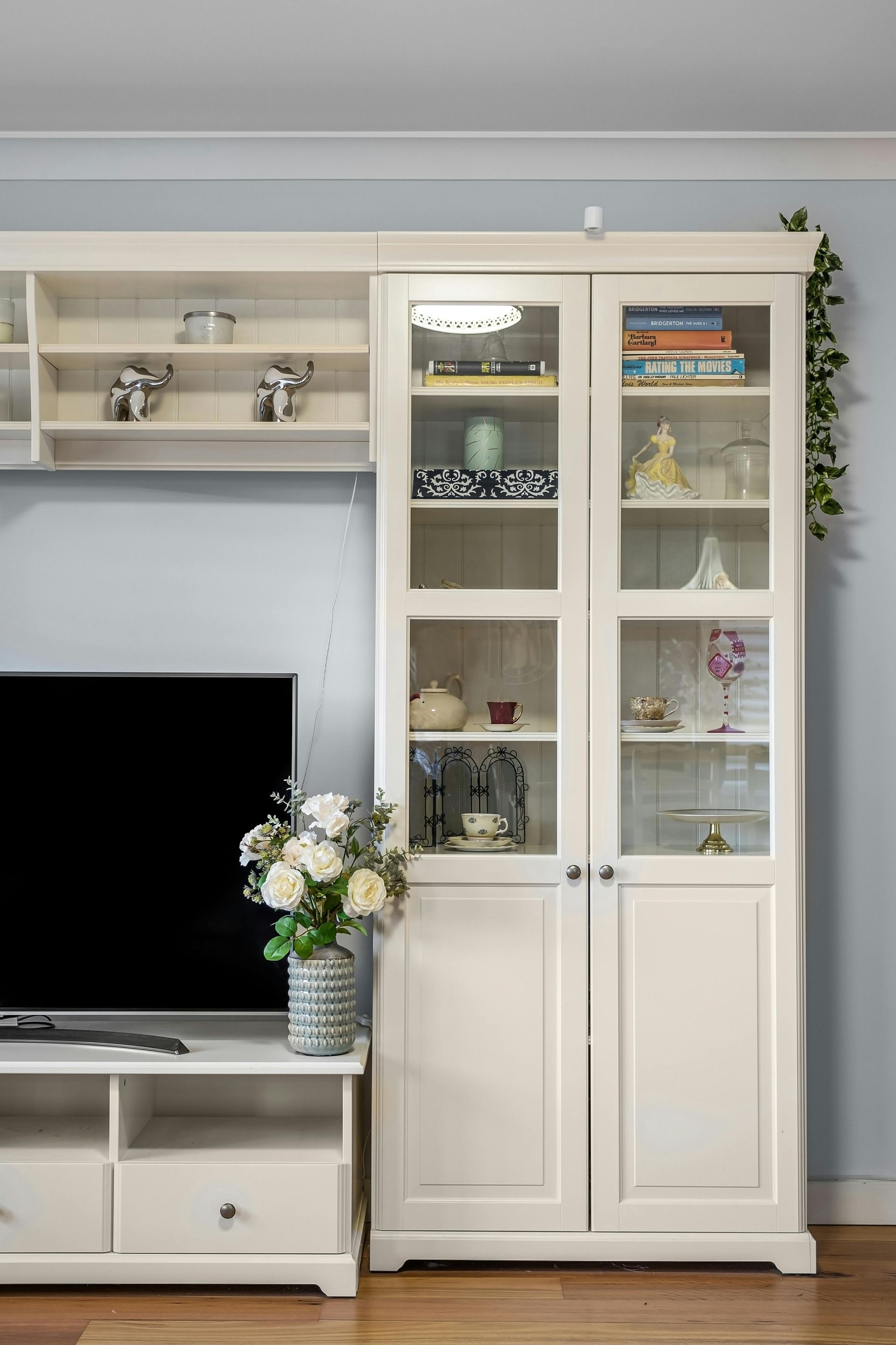 White display cabinet with glass doors beside a TV console in a bright living room